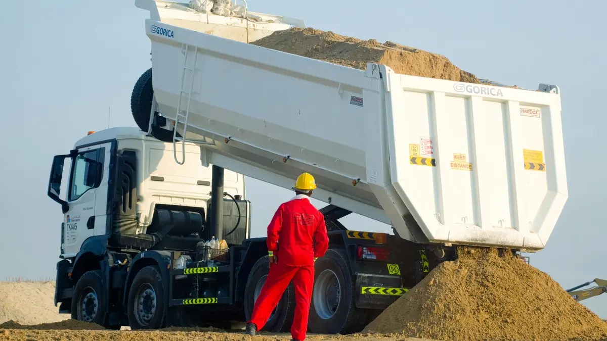 Tipper truck with its bed raised, discharging sand beside a site supervisor